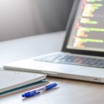 Open laptop with programming code on screen next to a notebook and pen on a desk