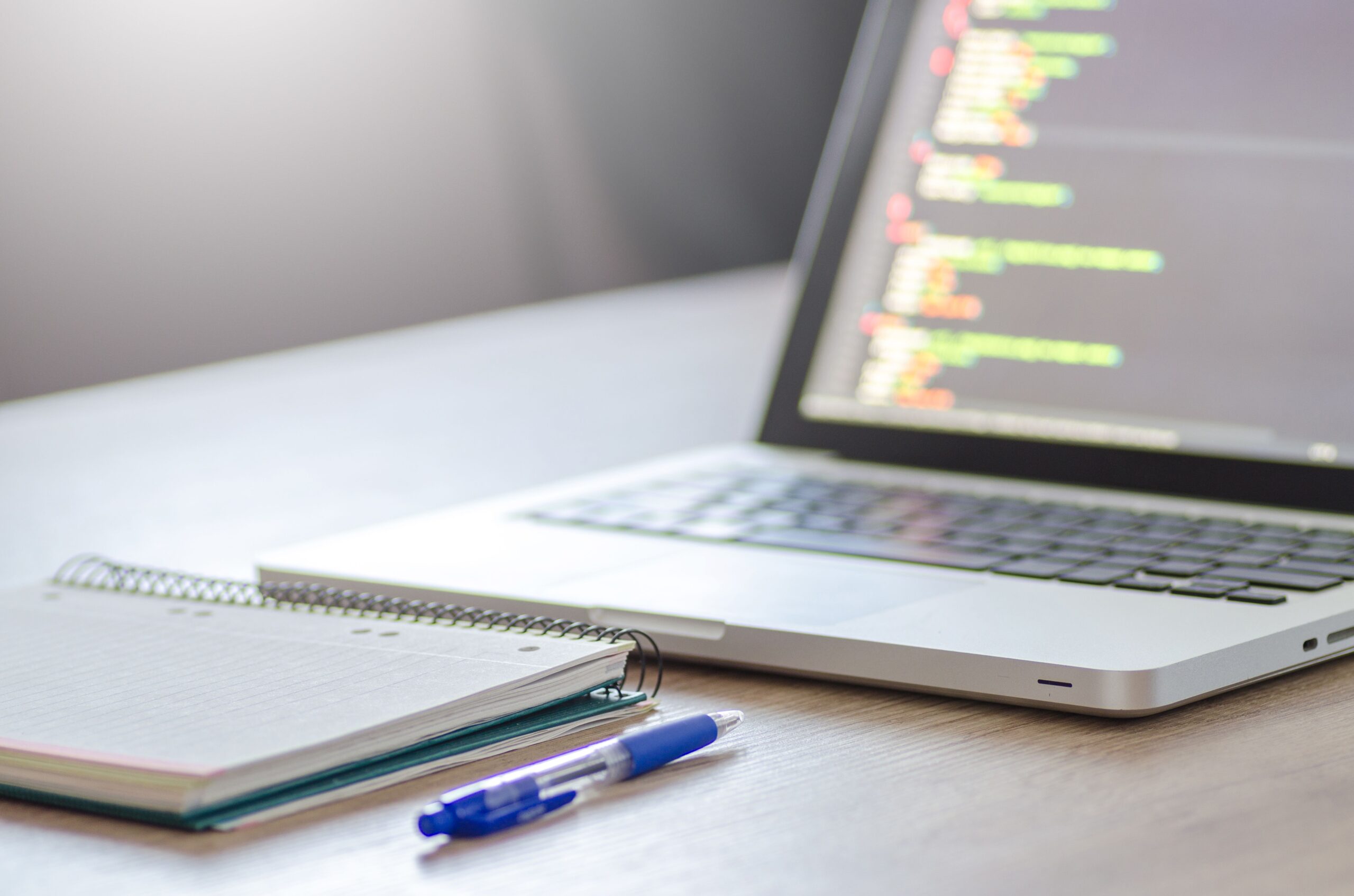 Open laptop with programming code on screen next to a notebook and pen on a desk